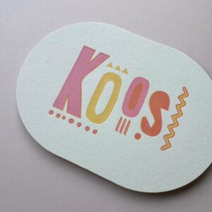 Een ovaal geboortekaartje met de naam Koos in vrolijke, versprongen letters. Letterpress gedrukt in 3 kleuren; roze, oranje, geel.